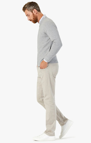 A man with short brown hair and a beard wears a light gray sweater, white sneakers, and 34 HERITAGE - COURAGE STRAIGHT LEG PANTS IN DAWN TWILL. He stands with his back to the camera, turning his head left against a plain white background.
