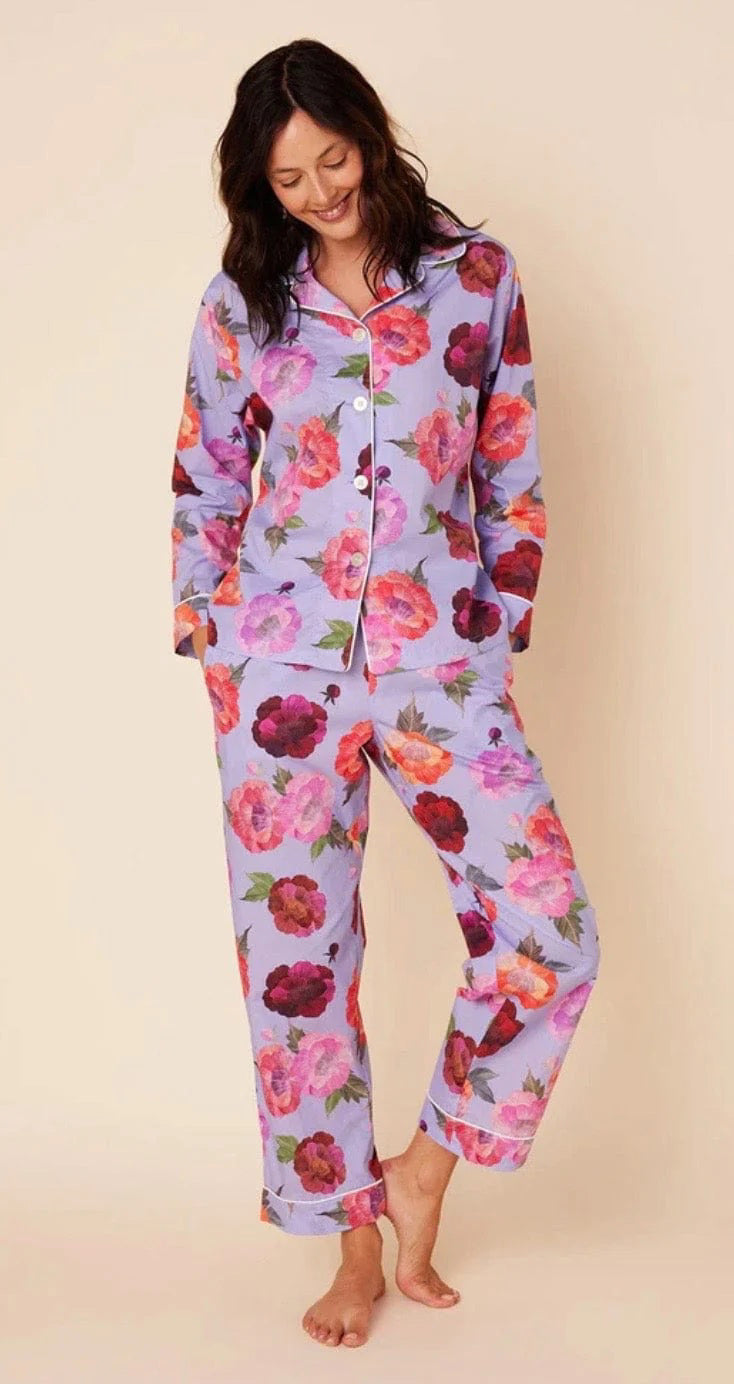 A woman is standing against a plain background, smiling downwards with her hands in the pockets of her Cat&#39;s Pajamas - Lavender Floral Luxe Pima Cotton Pajamas. The pajama set from Cat&#39;s Pajamas features a vibrant floral pattern with large red, pink, and purple flowers. She is barefoot and appears relaxed.
