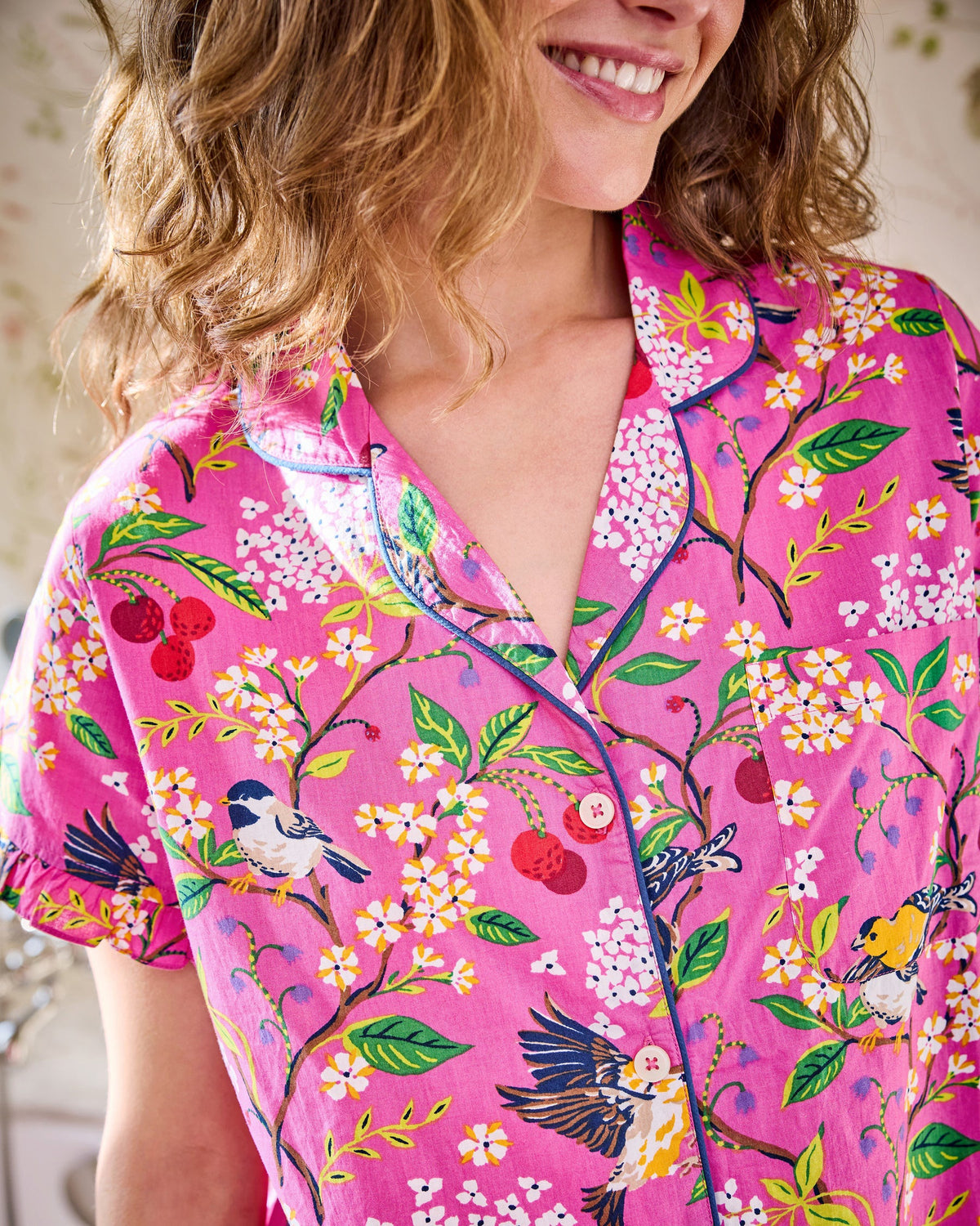 A woman wearing the PRINTFRESH Bird & Blossom Ruffle Pajama Short Set, featuring a pink, nature-inspired floral and bird print, stands against a plain light-colored wall with one hand in her shorts pocket, looking at the camera.