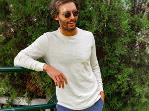 A man with short, curly hair and a beard wears the CRIQUET Cotton Slub Long Sleeve T-Shirt in Natural, styled with blue pants, standing against a plain white background.