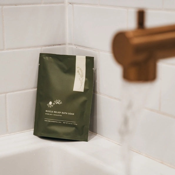 A green pouch of GIFTEN MARKET MUSCLE RELIEF BATH SOAK - EUCALYPTUS OIL rests on the edge of a bathtub lined with white tile, as water pours from a gold faucet in the foreground. Formulated with soothing eucalyptus oil and Epsom salt, this bath soak is an ideal choice for unwinding after a long day or adding to a thoughtful gift basket.