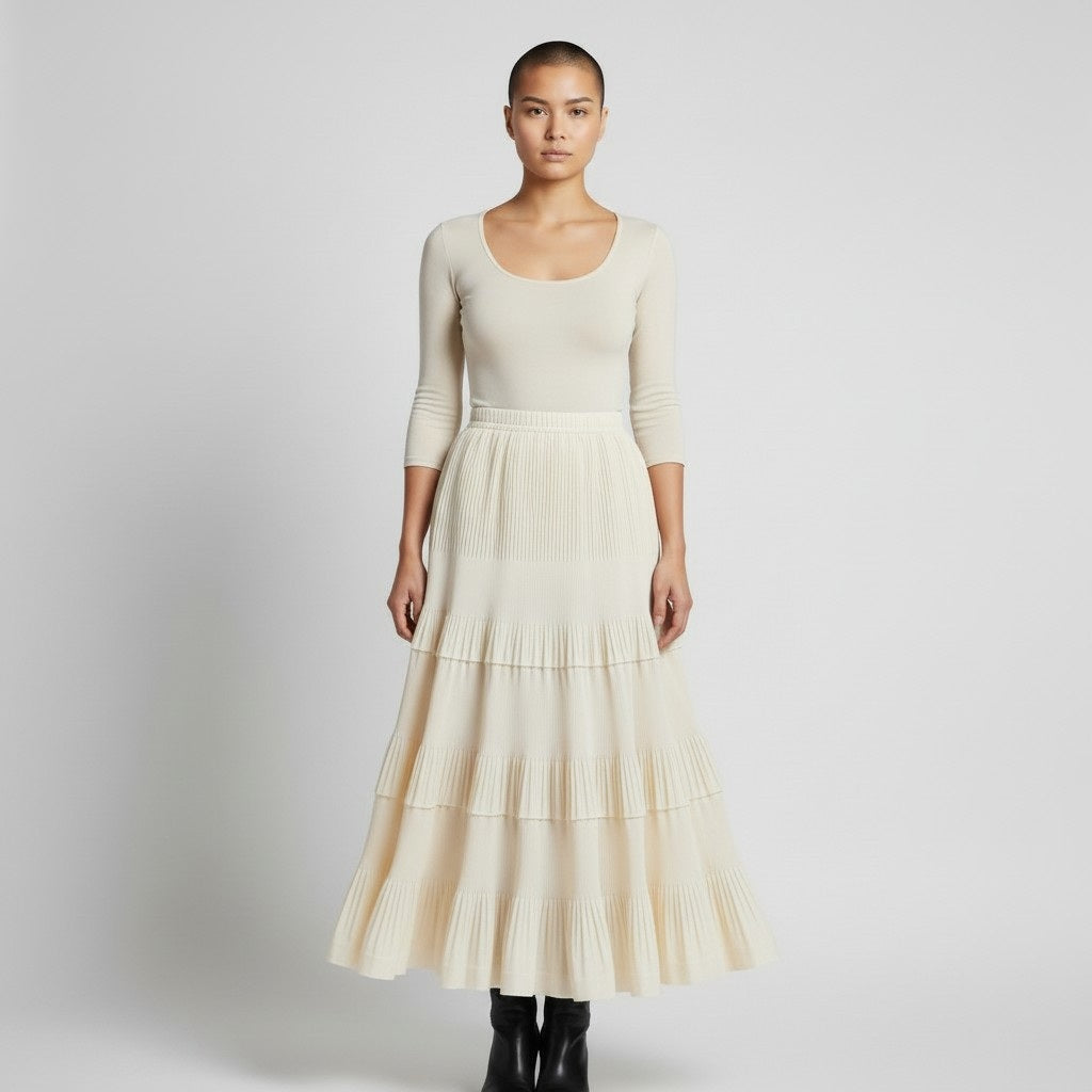 A person with a shaved head wears the MEIMEIJ RUFFLE SKIRT IN ANISE by MEIMEIJ and a fitted, cream long-sleeve top, standing against a plain light gray background.