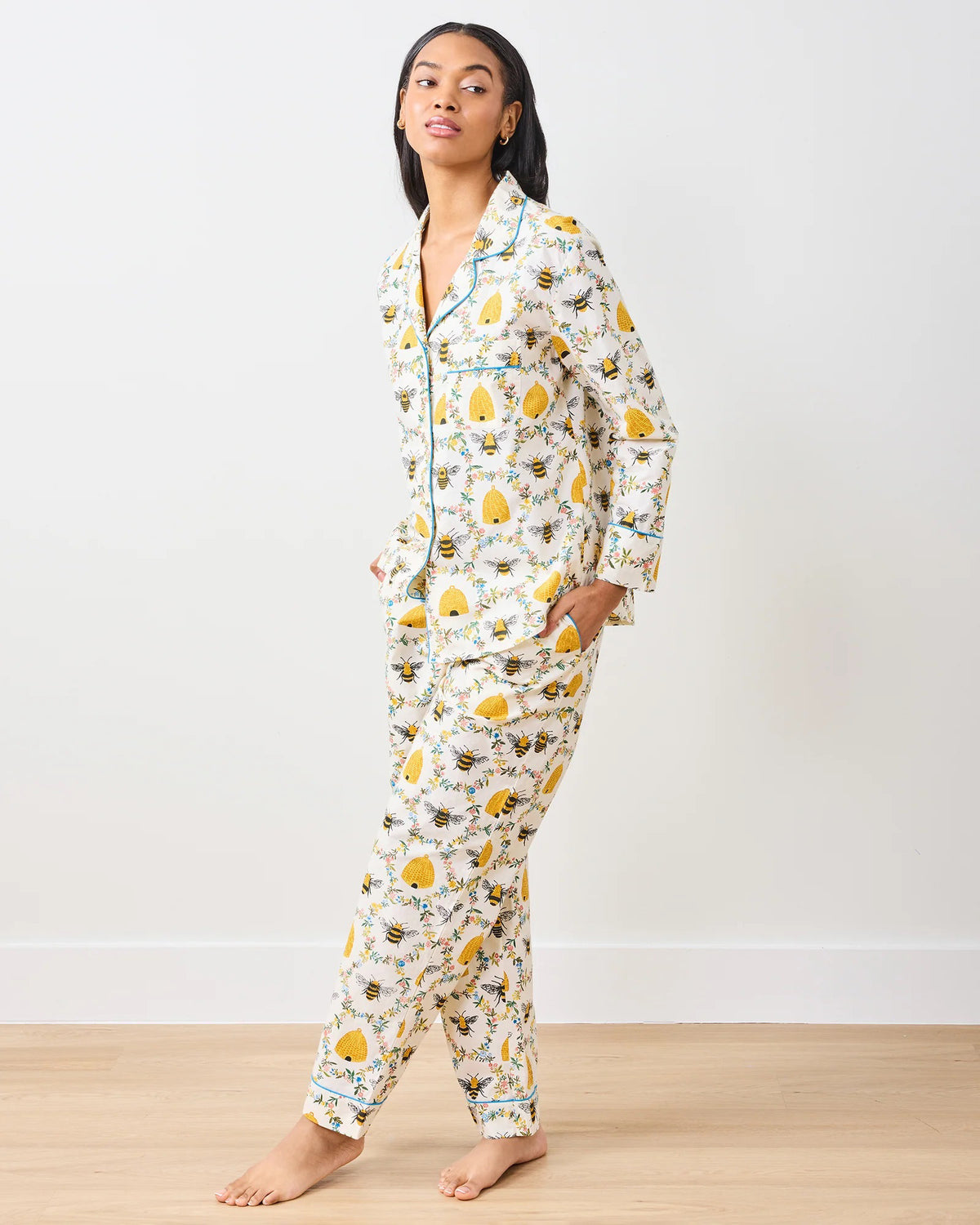A person stands barefoot on a wooden floor wearing the PRINTFRESH - SWEET BEE COTTON PAJAMA SET. This organic cotton pajama is white with yellow sun and cloud patterns, accented by black piping, resembling bee pajamas against a plain white wall.