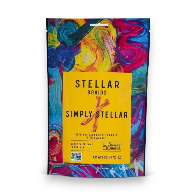 A vibrant bag of STELLAR SNACKS - SIMPLY STELLAR PRETZEL BRAIDS 5 OZ rests beside a white bowl filled with pretzel braids, set on a wooden board with a beige cloth nearby, all arranged on a white surface.