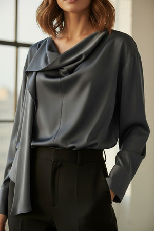 An elegantly dressed woman stands indoors by large windows, wearing the ELEH - BLOUSE WITH COWL NECK AND ATTACHED SCARF by ELEH—a silky grey blouse featuring a graceful draped neckline—paired with black tailored pants and black heels. Made in Italy, her outfit harmonizes beautifully with the room’s white walls and abundant natural light.