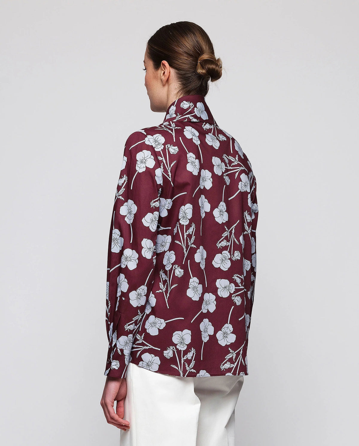 A woman stands by a window in a well-lit modern room, wearing the MIRTO 1956 Floral Print Blouse with Tie Neck in burgundy and high-waisted white pants, her hands casually tucked into her pockets.