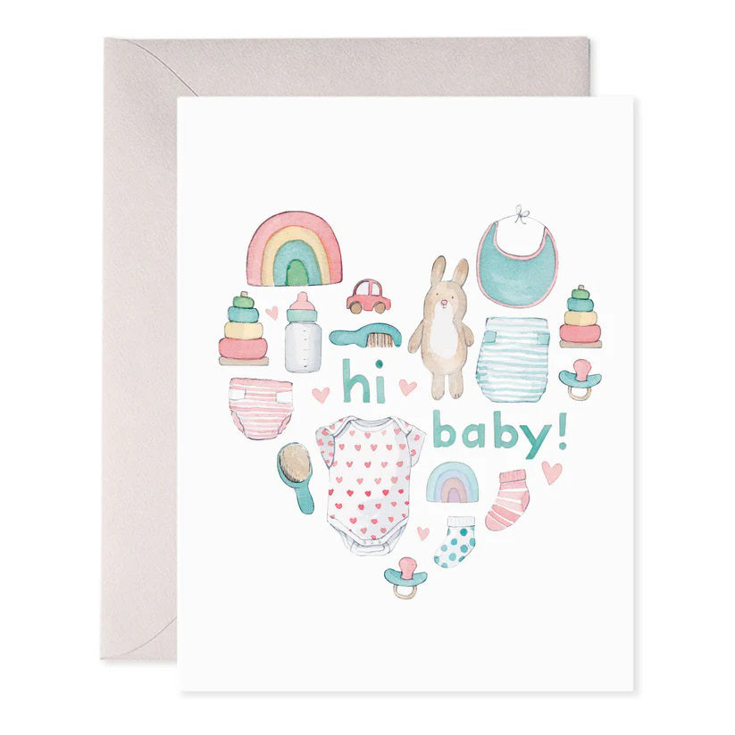 The E. FRANCES PAPER - BABY HEART GREETING CARD, by E. FRANCES PAPER, is handcrafted in the USA, featuring baby items like a rainbow, bib, bottle, bunny toy, onesie, stacking toys, pacifiers, and a wooden brush arranged in a heart shape with "hi baby!" at the center.