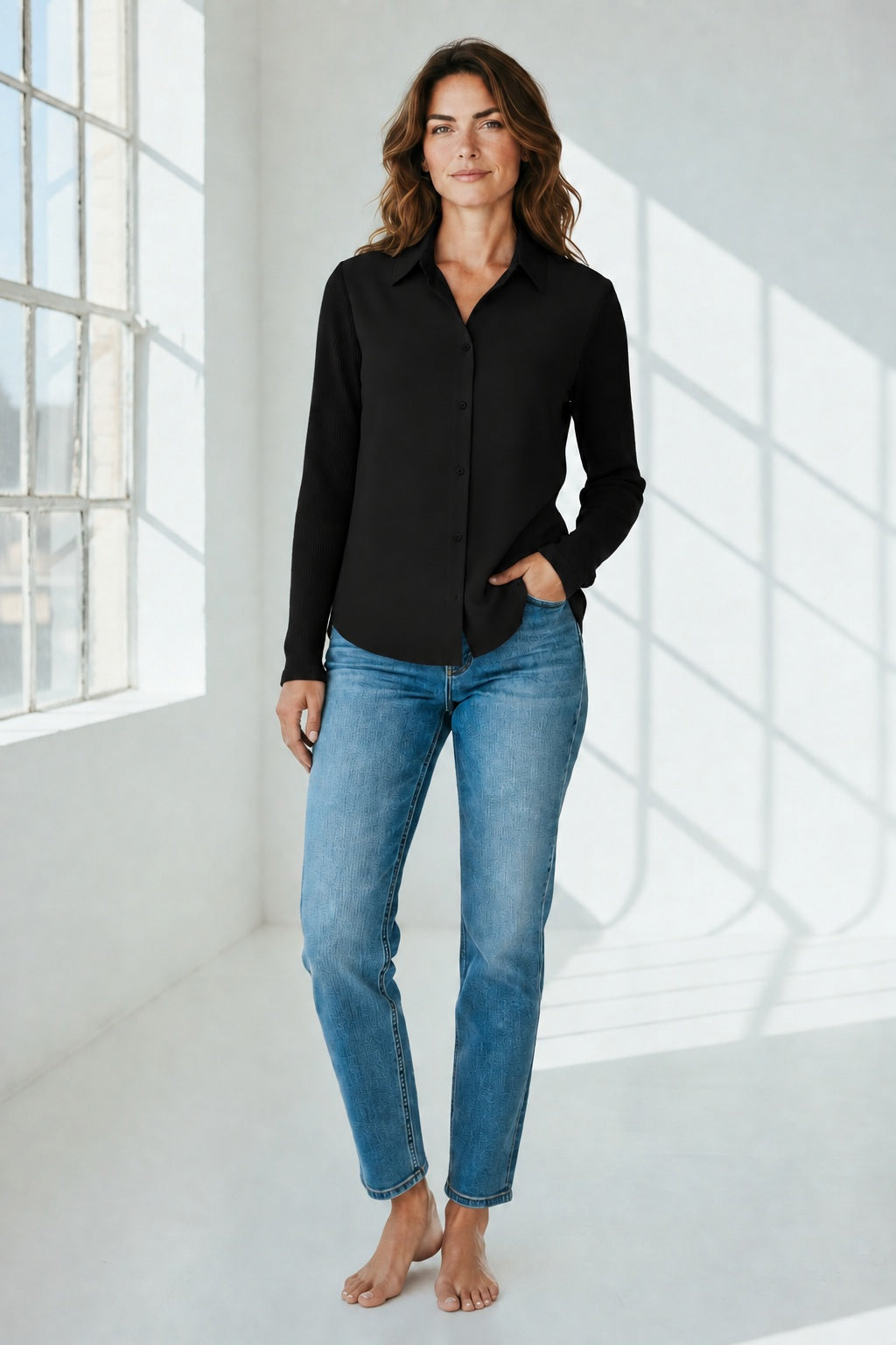 A woman stands barefoot in a bright room, wearing the SEVENTY VENEZIA button-up shirt with modal sleeves in black by SEVENTY and blue jeans, one hand in her pocket as natural light streams through large windows.