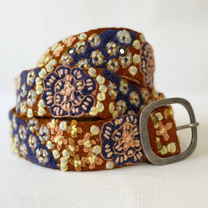 A close-up of a person in a blue denim shirt, dark pants, and the JENNY KRAUSS BLUE SKIES BELT, featuring vibrant floral embroidery and a large oval buckle—handcrafted in Peru.