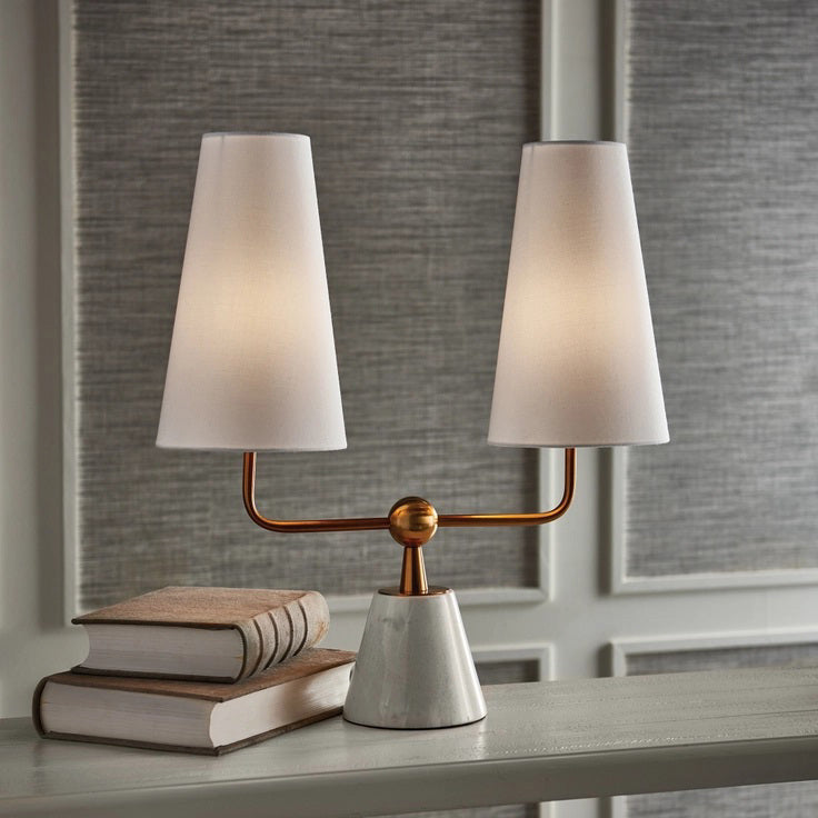 Introducing the Madison Dublet Lamp by NAPA HOME AND GARDEN, showcasing a modern design with two white conical shades on a gold bar. Its marble base and brass finish provide sophistication against a plain white background.