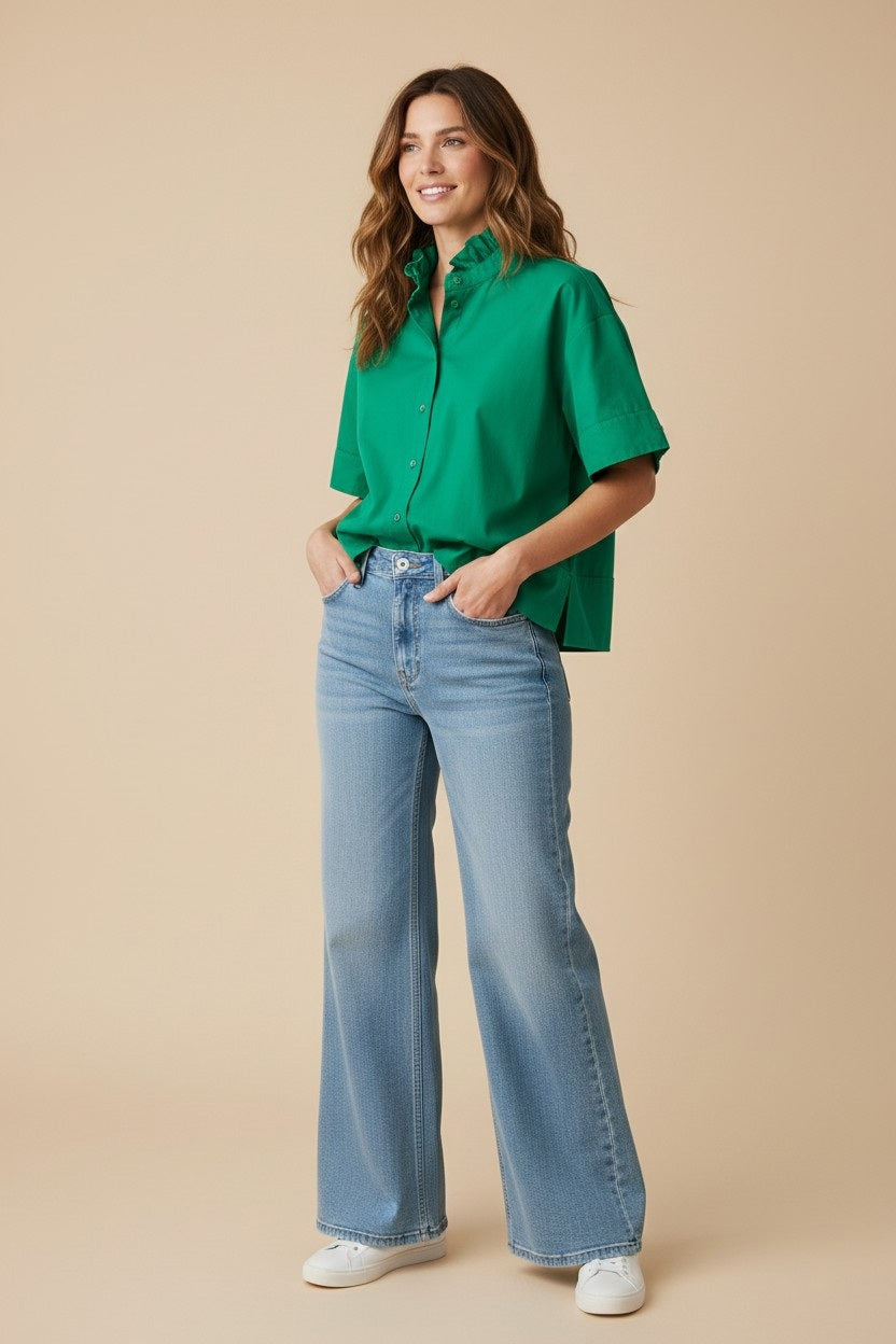 A woman smiles with hands in pockets, wearing the bright green KMJ - REESE SHIRT, light blue high-waisted wide-leg jeans, and white sneakers against a beige background.