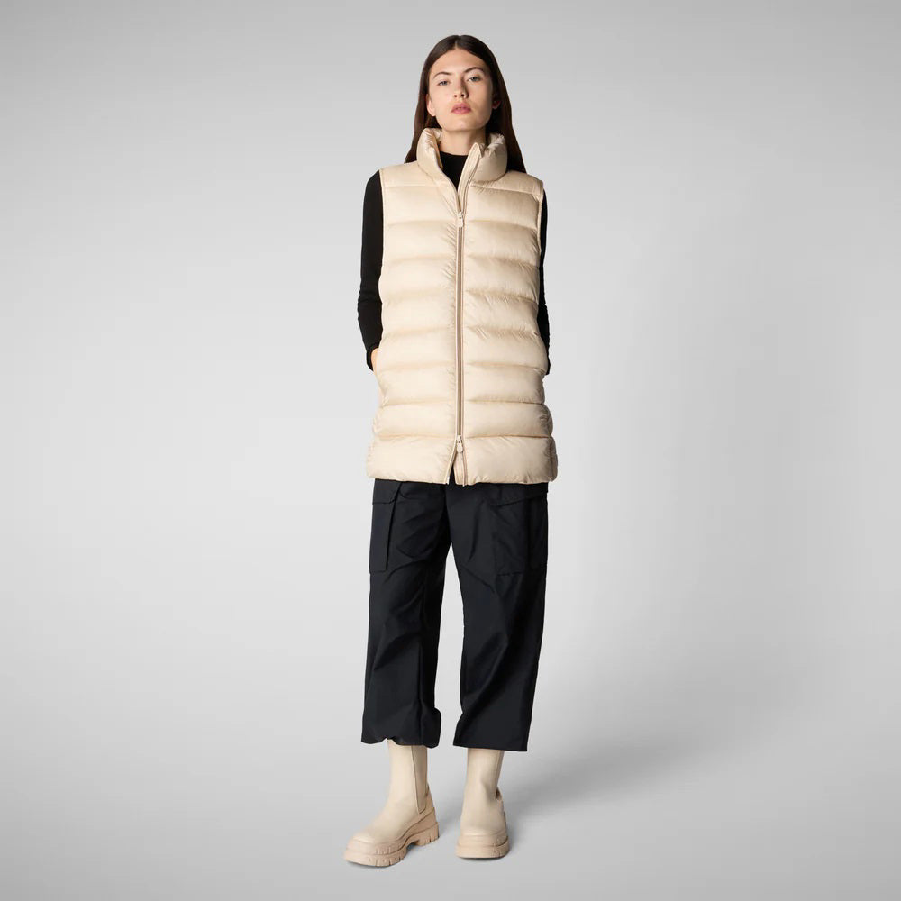 A person wearing a black cap, sunglasses, the SAVE THE DUCK - CORAL PUFFER VEST with animal-free down from SAVE THE DUCK over a black long-sleeve top, beige wide-legged pants, and black ankle boots stands against a plain background. The outfit appears to be styled for colder weather.