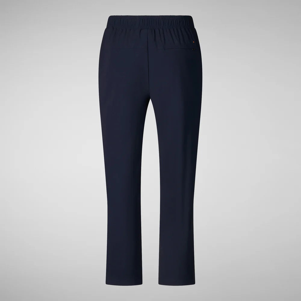 A person with shoulder-length hair and a neutral expression wears a white long-sleeve shirt, SAVE THE DUCK MILAN PULL ON PANT in black wrinkle-free fabric, and black slip-on shoes. Their hands rest at their sides against a plain background.