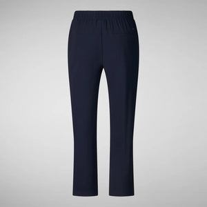 A person with shoulder-length hair and a neutral expression wears a white long-sleeve shirt, SAVE THE DUCK MILAN PULL ON PANT in black wrinkle-free fabric, and black slip-on shoes. Their hands rest at their sides against a plain background.