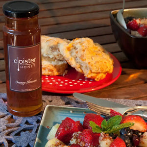 A tall, clear glass jar filled with dark amber honey labeled "Cloister Honey" with "Orange Blossom" beneath it on a brown label. The jar has a black metal lid and is photographed against a white background, highlighting the rich color and subtle citrus undertone of the natural Cloister Honey - Orange Blossom Honey 12oz.