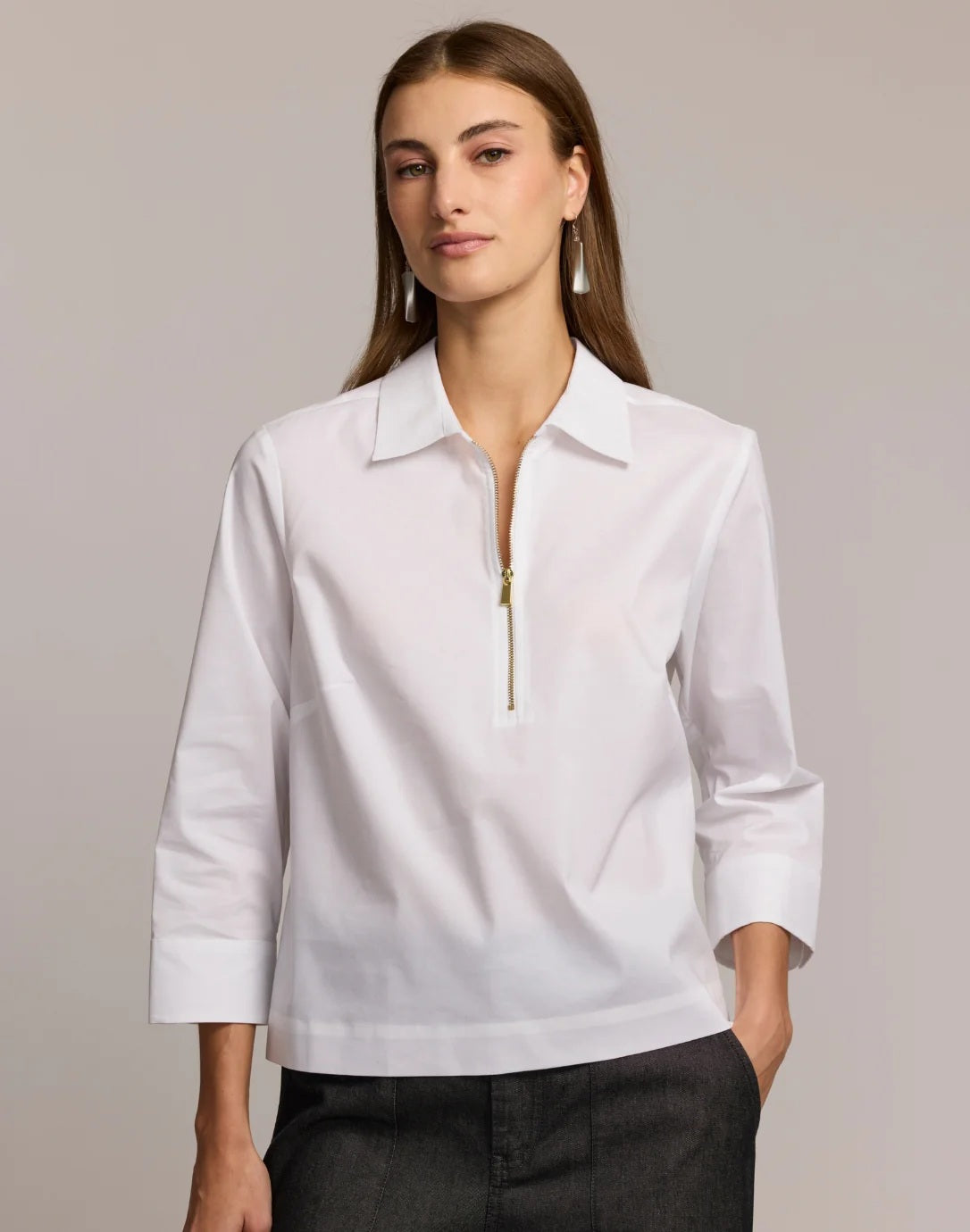 A woman stands against a plain, light background wearing the HINSON WU Alessa 3/4 Sleeve Shirt in stretch polished cotton—a crisp white blouse with a collared neckline and partial front zipper—paired with dark pants.