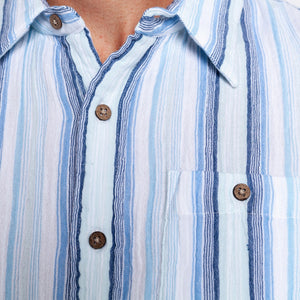 A man wearing the CRIQUET - THE BEACHCOMBER SHIRT IN BLUE by CRIQUET, paired with light beige shorts, stands against a plain white background, looking to his left with one hand in his pocket.