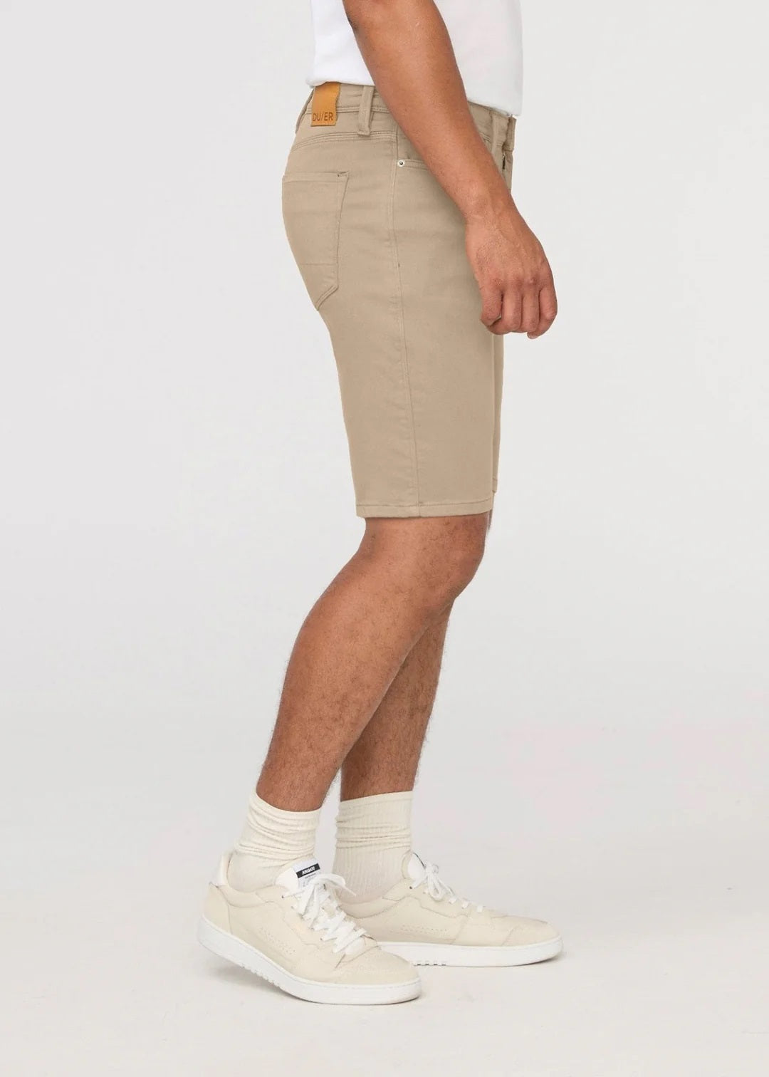 A person wearing a white t-shirt, DUER No Sweat Relaxed Shorts in Desert Khaki, white crew socks, white sneakers, and a wristwatch stands against a plain light background.