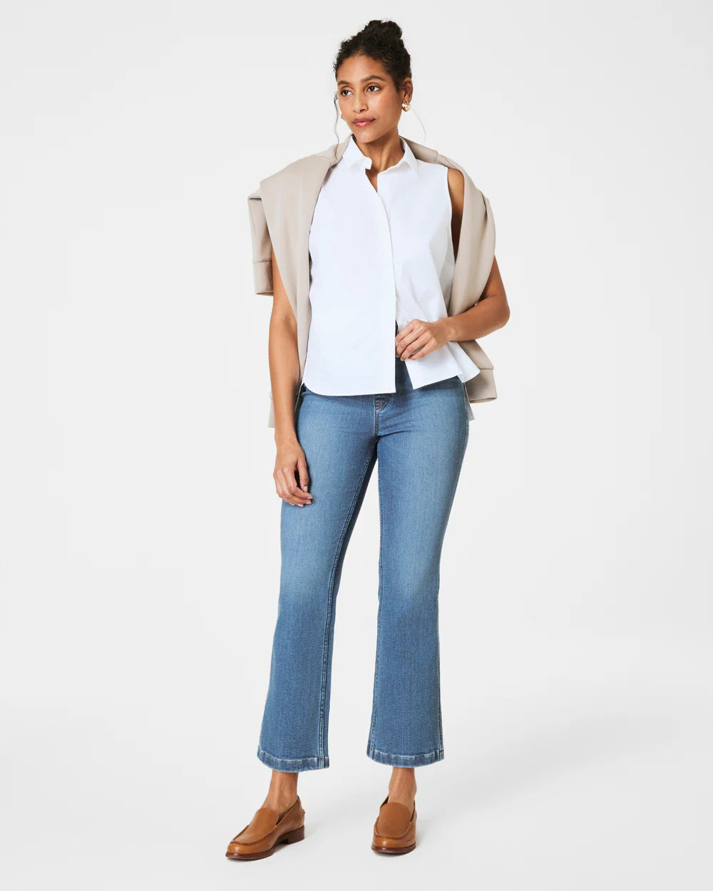 A person poses against a plain backdrop, wearing a sleeveless white blouse and SPANX Pull On Kick Flare Jeans with brown loafers. A beige jacket is draped over their shoulders, and their hair is styled in a bun.