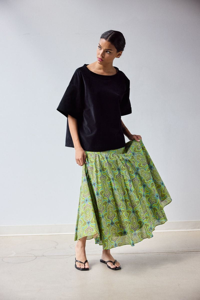 A woman stands indoors in a loose black top and the HOPE FOR FLOWERS - CIRCLE SKIRT IN GREEN FOULARD, made from soft organic cotton lawn. She holds out one side of her GOTS certified skirt by HOPE FOR FLOWERS, paired with black sandals.