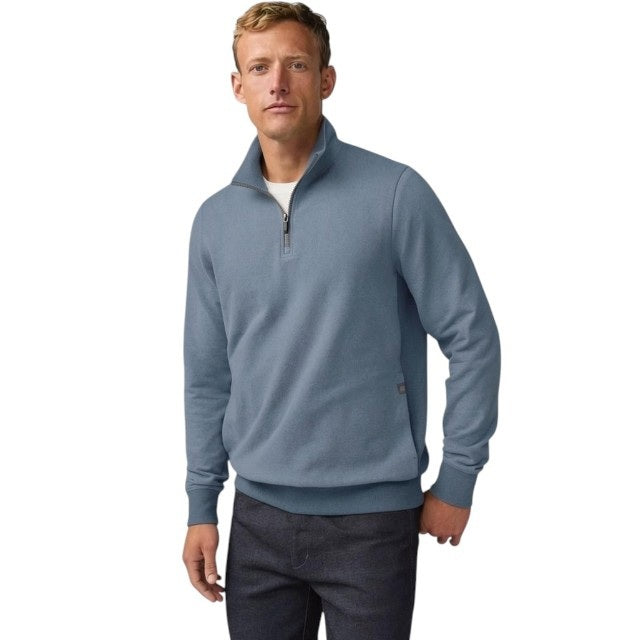 A man with short light brown hair stands in profile, wearing the MACK WELDON Ace 1/2 Zip Sweater in Herringbone and dark pants, one hand in his pocket, against a plain light gray background.