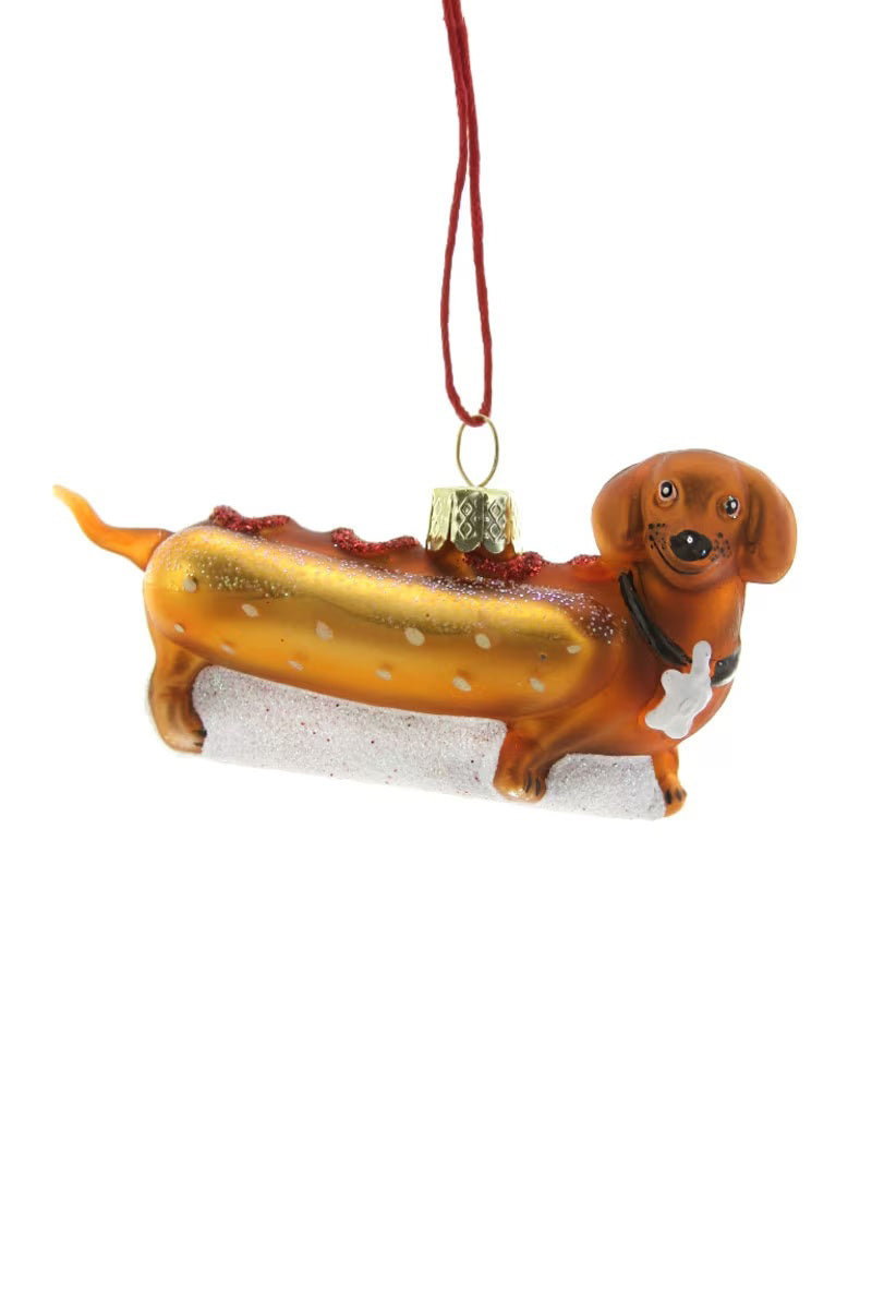 The CODY FOSTER WIENER PUP DOG ORNAMENT is a 3.25&quot; glass dachshund-shaped Christmas ornament, styled like a hot dog in a bun and hanging by a red string—a whimsical addition to your holiday decor.