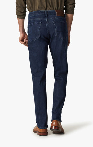 A man in a green long-sleeve shirt, brown shoes, and 34 HERITAGE Charisma Relaxed Straight Leg Jean in Dark Midnight Brushed Urban wash stands with his back to the camera against a plain white background.