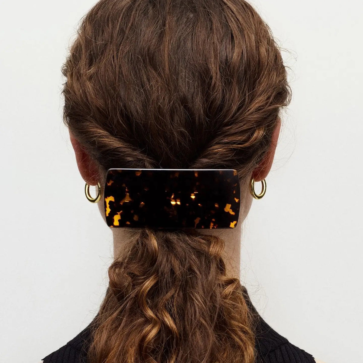A person with brown wavy hair in a low twist, secured by the MACHETE - JUMBO BOX BARRETTE IN TORTOISE, is seen from behind wearing gold hoop earrings and a black top.