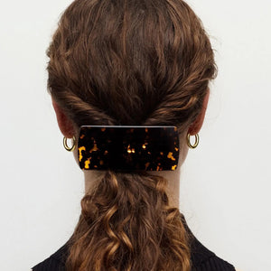 A person with brown wavy hair in a low twist, secured by the MACHETE - JUMBO BOX BARRETTE IN TORTOISE, is seen from behind wearing gold hoop earrings and a black top.