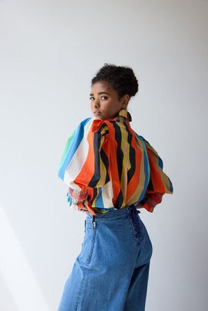 A person with curly hair wears the HOPE FOR FLOWERS Smocked Shirt in Awning Stripes, featuring puffed sleeves and a high collar, paired with high-waisted blue jeans against a plain, light background.