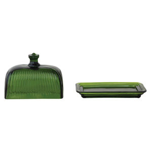The CREATIVE COOP GREEN PRESSED GLASS BUTTER DISH is displayed with green glass containers and bottles on a textured white surface, beside a folded towel and a partially visible book.