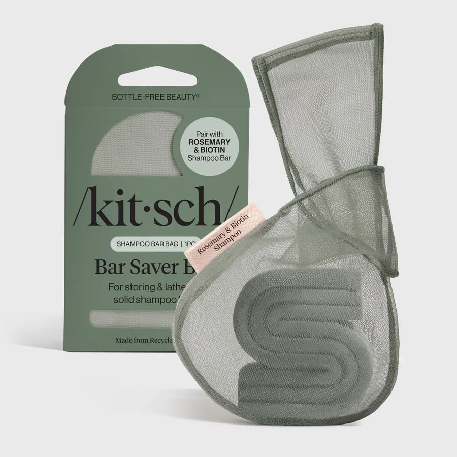 A green, eco-friendly mesh bag from KITSCH sits beside green packaging labeled “KITSCH - ROSEMARY & BIOTIN SHAMPOO BAR SAVER BAG,” designed for storing solid shampoo bars and extending their lifespan. A pink tag reads “Rosemary & Biotin Shampoo Bar.” Both the bag and box are displayed against a white background.