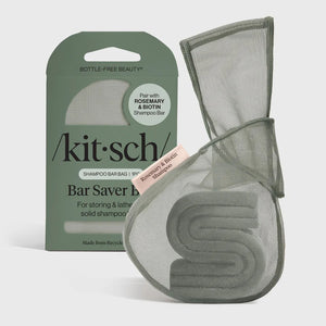 A green, eco-friendly mesh bag from KITSCH sits beside green packaging labeled “KITSCH - ROSEMARY & BIOTIN SHAMPOO BAR SAVER BAG,” designed for storing solid shampoo bars and extending their lifespan. A pink tag reads “Rosemary & Biotin Shampoo Bar.” Both the bag and box are displayed against a white background.