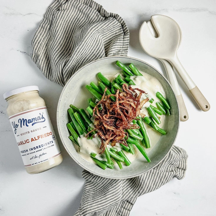 A jar of YO MAMA'S - ROASTED GARLIC ALFREDO by YO MAMA'S FOODS rests on an orange background, featuring a 16 oz (453g) label that boasts "Fresh Ingredients," "No Sugar Added," "Gluten-Free," with roasted garlic and parmesan cheese.