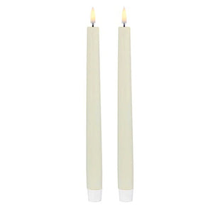 Two PIFFANY UYUNI Ivory Slim Taper Candles (1x12.6) with small yellow flames stand upright side by side against a plain white background, exuding timeless elegance.