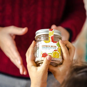 A clear mason jar with a silver lid containing colorful, citrus-flavored hard candies. The label reads "BUTTERFIELDS - CITRUS BUDS" and "Tangy, Zesty, Juicy Fruits" with illustrations of oranges, lemons, and grapefruits. The jar's net weight is 7.3 oz (204 g).