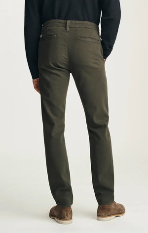 A person stands with their back to the camera against a neutral background, wearing the 34 HERITAGE - VERONA Straight Leg Chino Pant in Jasper High Flyer paired with a black long-sleeve shirt and brown suede shoes.