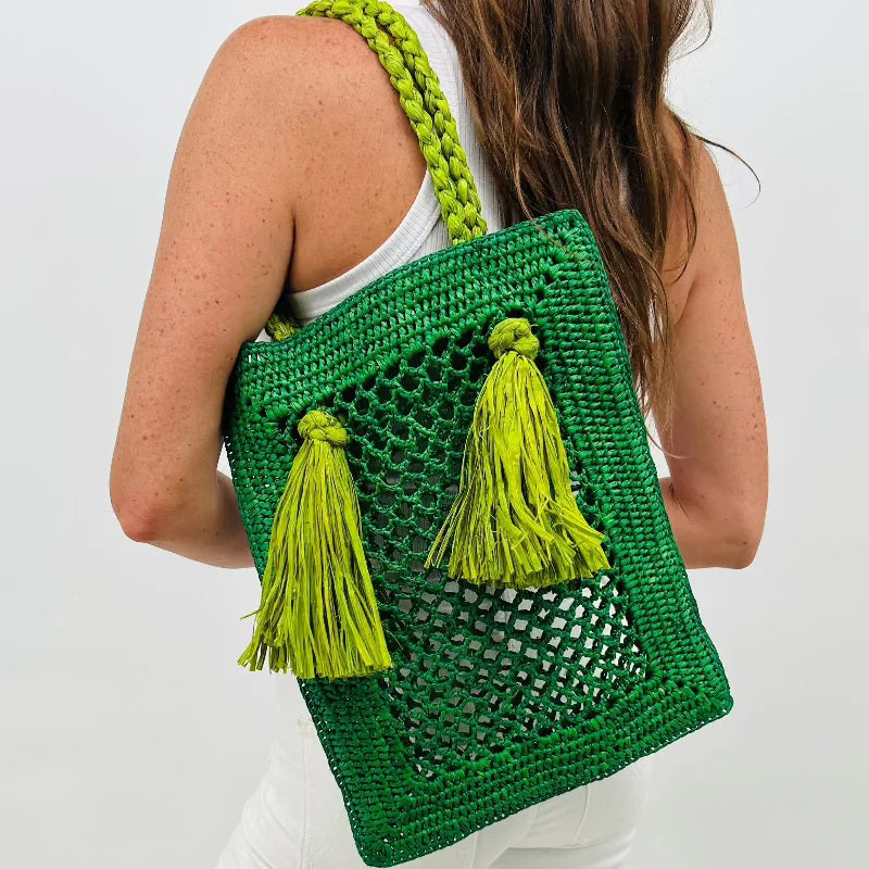 A woman in white is seen from behind holding the BALDIZ - JUNIPER RAFFIA BAG IN EMERALD AND LIME, featuring two large yellow-green tassels and thick braided handles, against a plain white background.