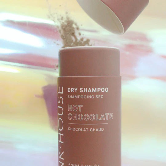 A cylindrical bottle of PINK HOUSE ORGANICS - DRY SHAMPOO HOT CHOCOLATE by PINK HOUSE ORGANICS is shown. The label highlights its 100% natural ingredients and promotes it as a quick and easy solution for greasy hair. The bottle is displayed against a pastel-colored, blurred background.