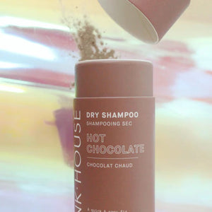 A cylindrical bottle of PINK HOUSE ORGANICS - DRY SHAMPOO HOT CHOCOLATE by PINK HOUSE ORGANICS is shown. The label highlights its 100% natural ingredients and promotes it as a quick and easy solution for greasy hair. The bottle is displayed against a pastel-colored, blurred background.