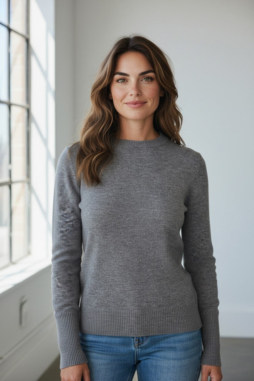A woman with long brown hair stands indoors near a large window, smiling softly at the camera while wearing the KINROSS CASHMERE Modern Crew Neck Cashmere Sweater in Granite and blue jeans.