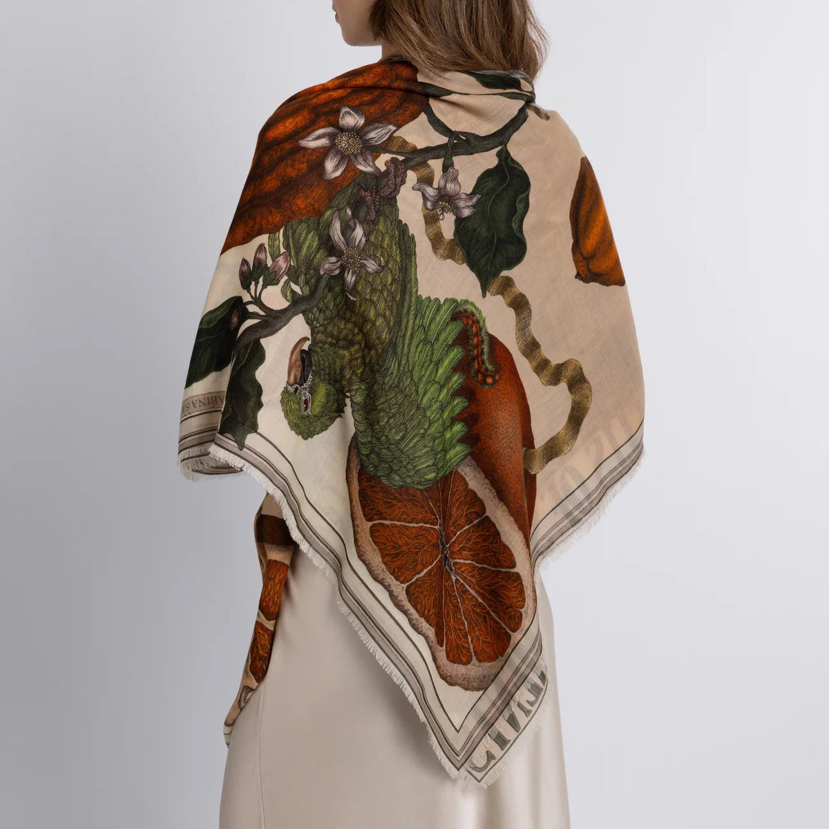 A person elegantly stands against a plain background wearing the SABINA SAVAGE ORANGES AND LEMONS 135CM cashmere scarf in Citrine/Zest, featuring animals, leaves, and objects that contrast beautifully with their light-colored dress.