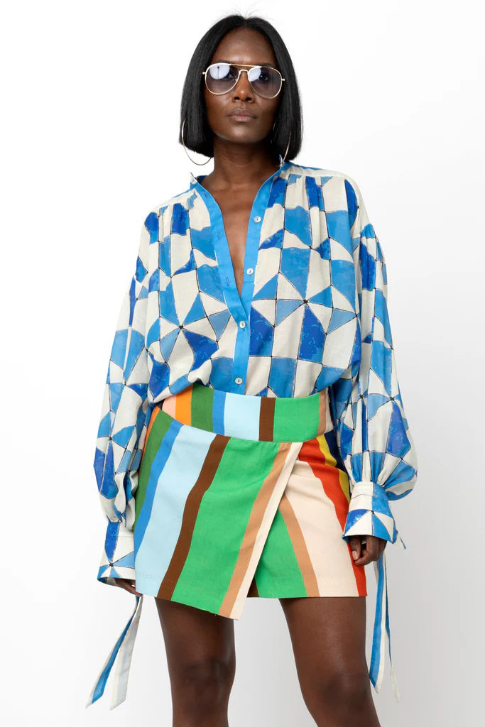 A person stands against a white background, wearing the HOPE FOR FLOWERS Wrist Tie Shirt in a blue and white geometric pattern, paired with a short, multicolored striped skirt. They have shoulder-length hair and are wearing sunglasses. Their hands rest by their sides.