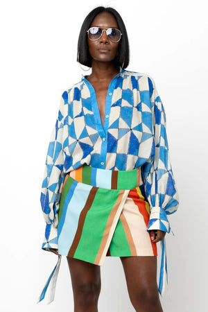 A person stands against a white background, wearing the HOPE FOR FLOWERS Wrist Tie Shirt in a blue and white geometric pattern, paired with a short, multicolored striped skirt. They have shoulder-length hair and are wearing sunglasses. Their hands rest by their sides.