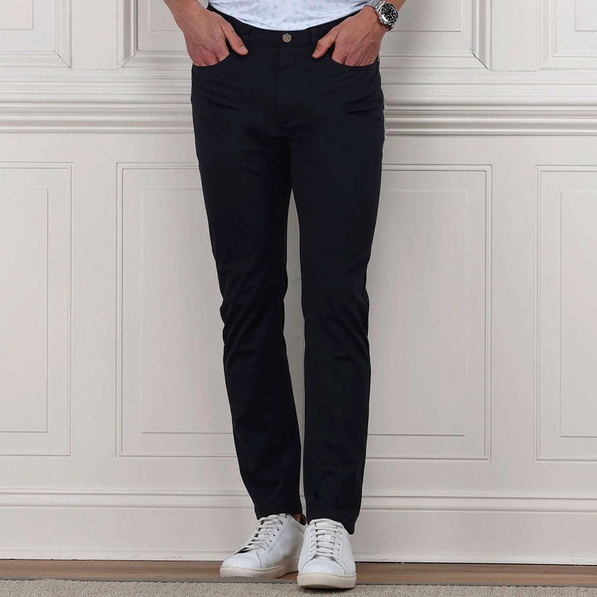 A man wearing a blue patterned polo, COLLARS AND CO Velocity Pull-On Performance Pants in Navy, and white sneakers stands indoors, confidently leaning on a golf bag against a white paneled wall.
