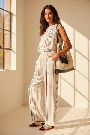 A person stands on white pebbles wearing ELEH’s STRIPED TROUSER—a loose, wide-leg design with a matching sleeveless top in a soft rayon blend—paired with brown sandals and a beige woven bag.