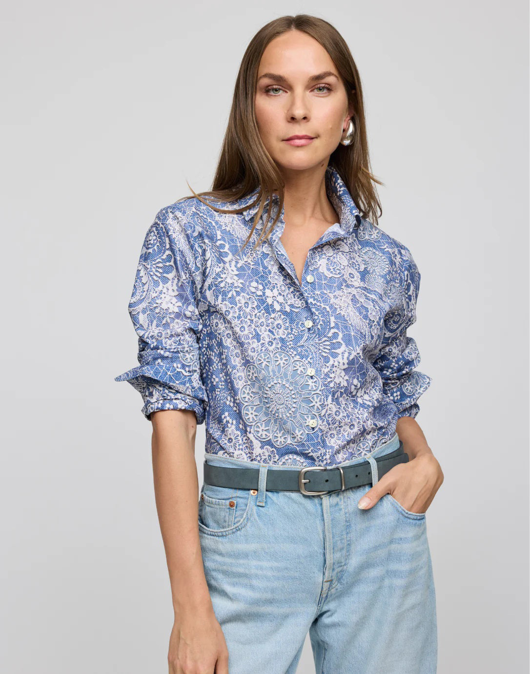 Someone with long hair poses against a plain background, wearing the HINSON WU - MARGOT LONG SLEEVE LACE PRINT SHIRT and light blue jeans. One hand is on their hip, the other by their side, exuding a breathable aura of comfort and style.
