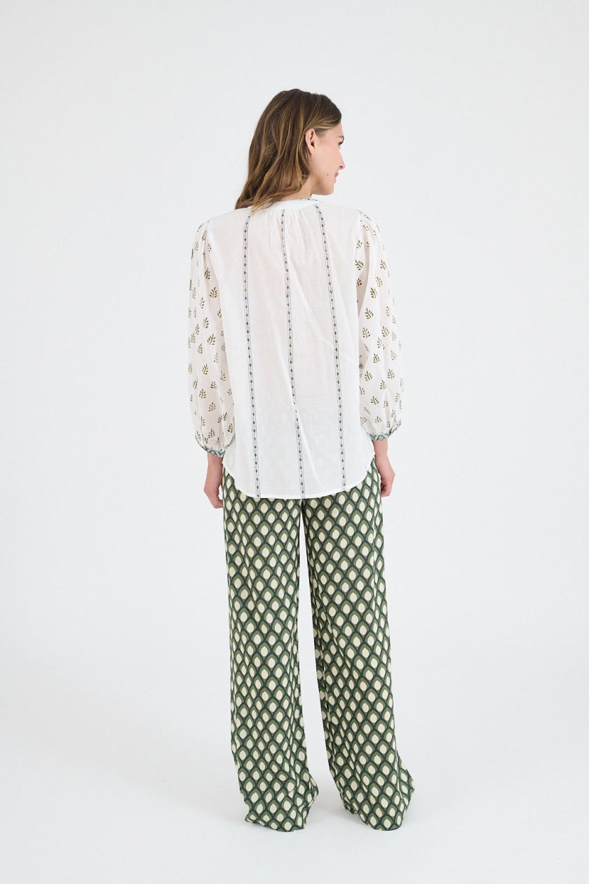 A woman wears the STORIATIPIC LUCIE FLOWER BLOUSE—a cotton blouse with patterned sleeves and green trim—paired with wide-leg green patterned pants, standing against a white background in sandals, evoking casual elegance.