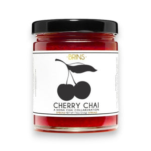 A glass jar of BRINS - CHERRY CHAI SPREAD & PRESERVE features a black lid and a label adorned with an illustration of two cherries alongside flavor details and collaboration notes. Made by BRINS with locally sourced New York sour cherries, this preserve is expertly infused with aromatic chai spices for a distinctive and flavorful experience.