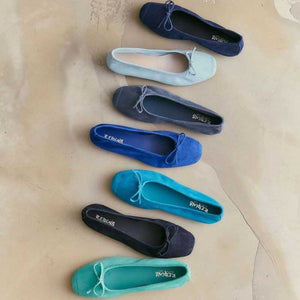 Someone stands on a light floor, one foot slightly raised, wearing REQINS - HARMONY suede ballerina flats in marine with a small bow, paired with blue jeans featuring frayed hems.